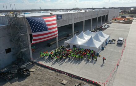 An aerial view of a building under construction. An American flag is attached to the side of the building, and dozens of construction workers in personal protective equipment are gathered in front of white event tents.